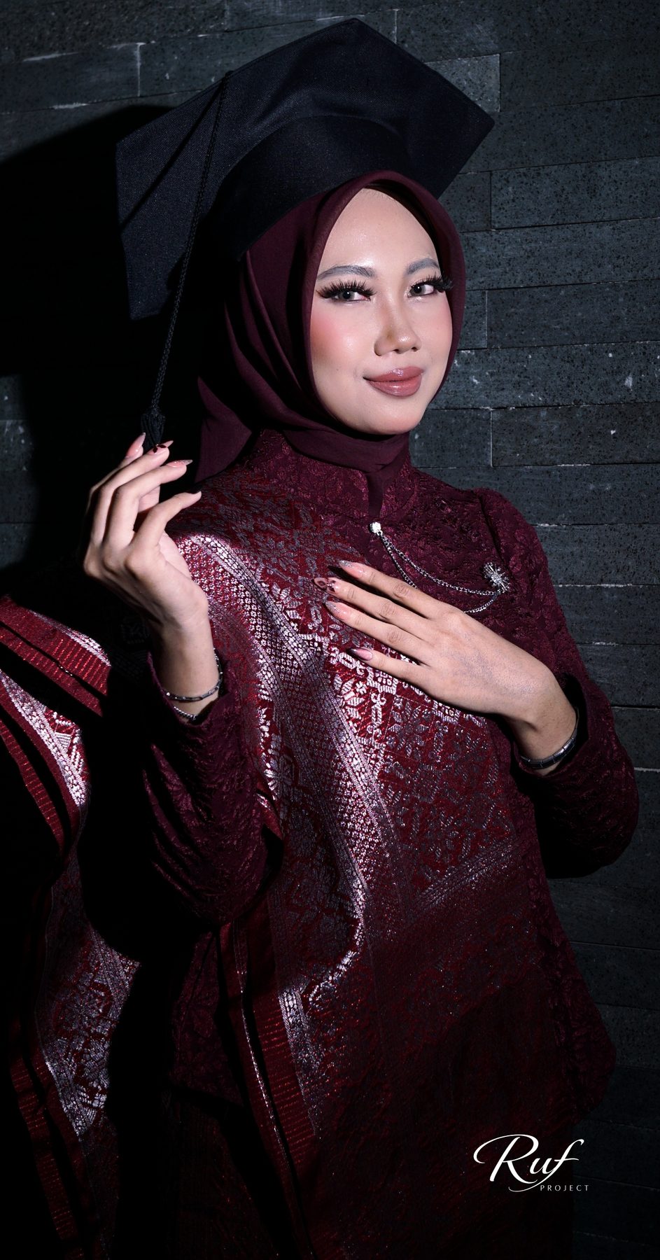 Wisudawati bertoga dengan songket marun berpose di depan dinding batu